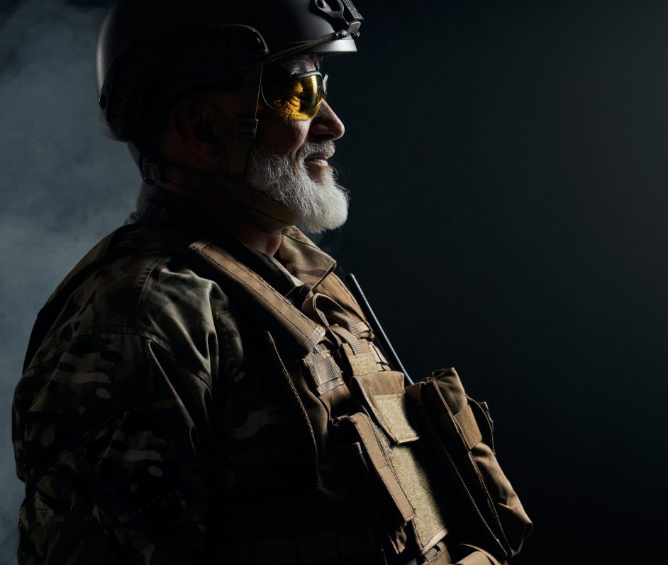 Side view of old military officer looking away. Crop portrait of bearded veteran in camouflage uniform, glasses and helmet posing in smoky dark atmosphere. Concept of military, army.